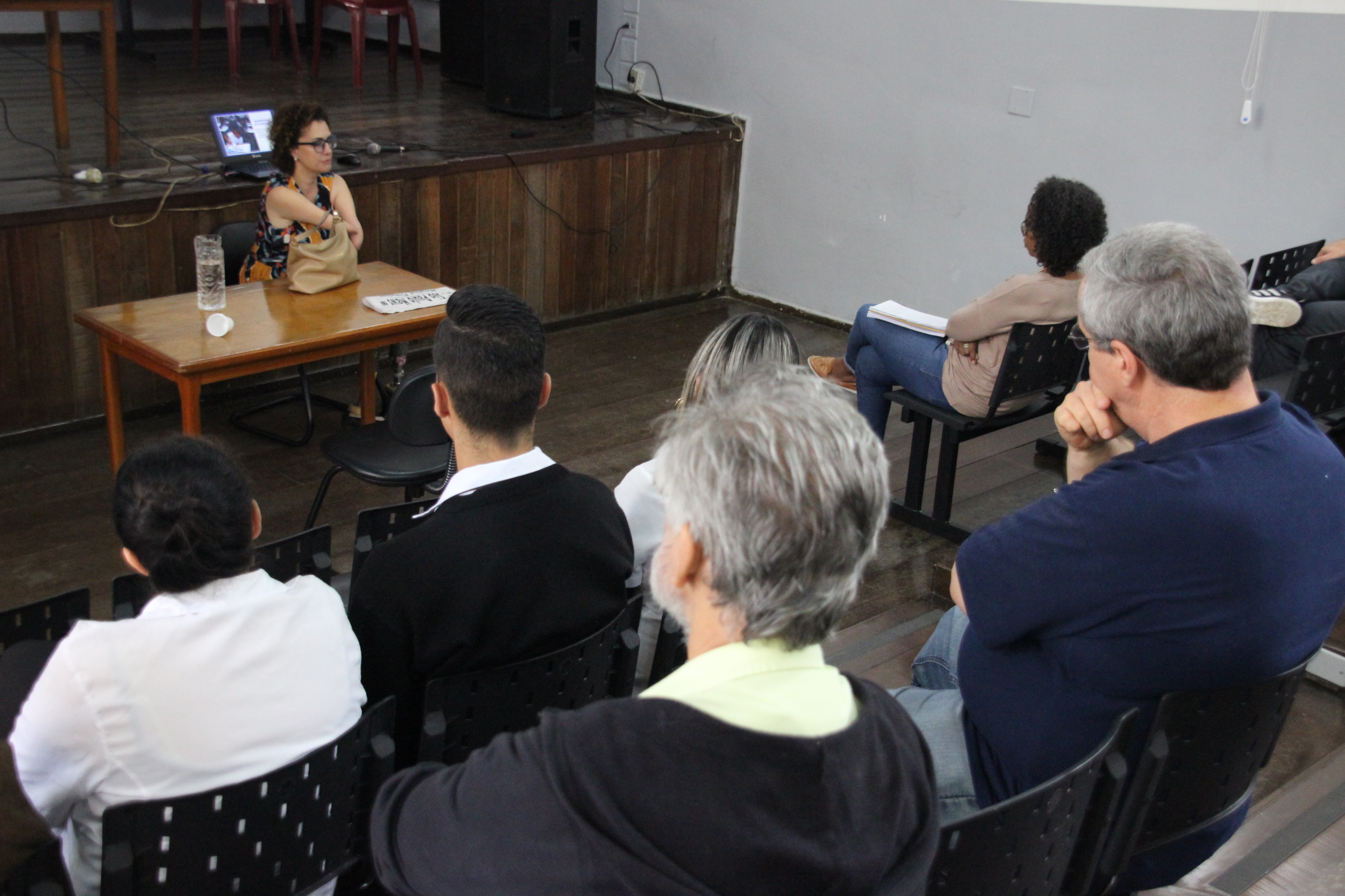 Imagem registrada de frente para a palestrante, da lateral esquerda para a direita com participantes de costas para a foto e a palestrante de frente. 